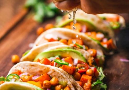 A hand is squeezing lime over a row of tacos filled with chickpeas, avocado, cilantro, and diced vegetables on a wooden surface.