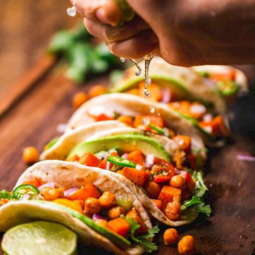A hand is squeezing lime over a row of tacos filled with chickpeas, avocado, cilantro, and diced vegetables on a wooden surface.