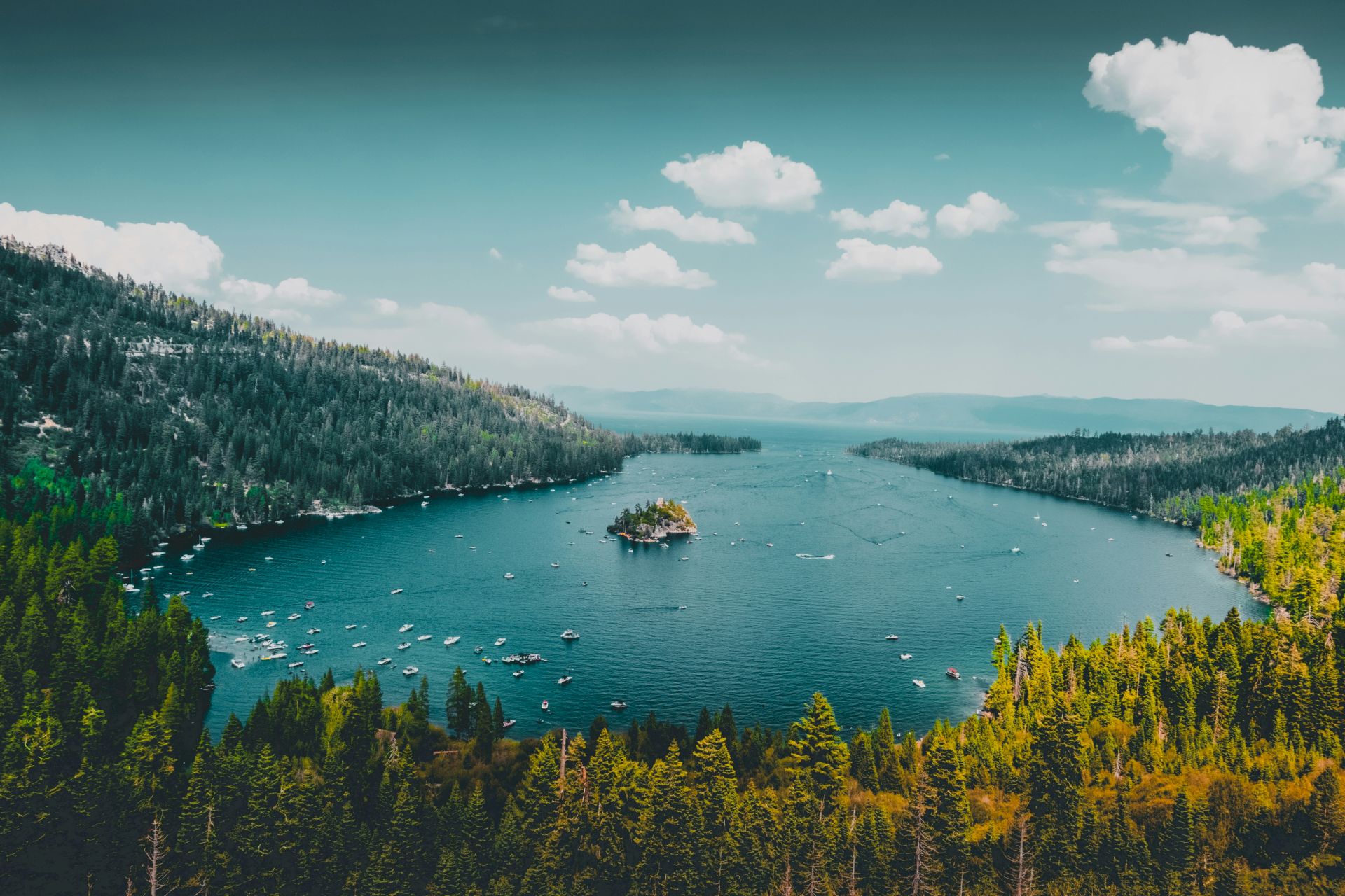 A scenic lake surrounded by lush green forests and mountains, featuring a small island in the center with numerous boats dotting the water.