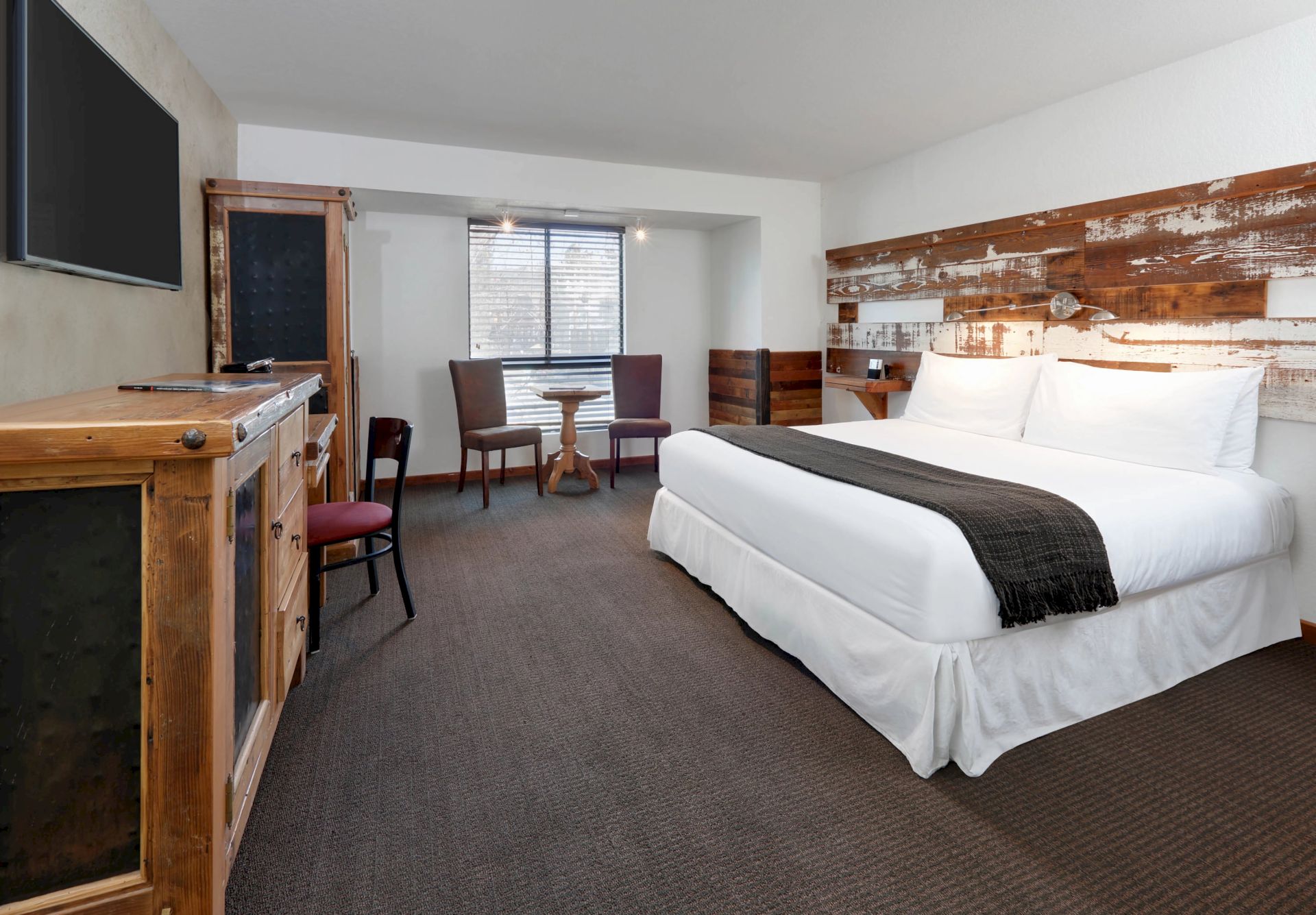 The image shows a cozy hotel room with a king-sized bed, rustic decor, a TV, chairs, and a round table near a window, creating a welcoming space.