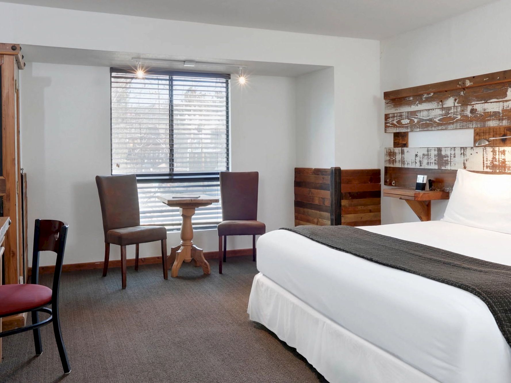 A hotel room with a bed, two chairs, a small table, and a window with blinds.