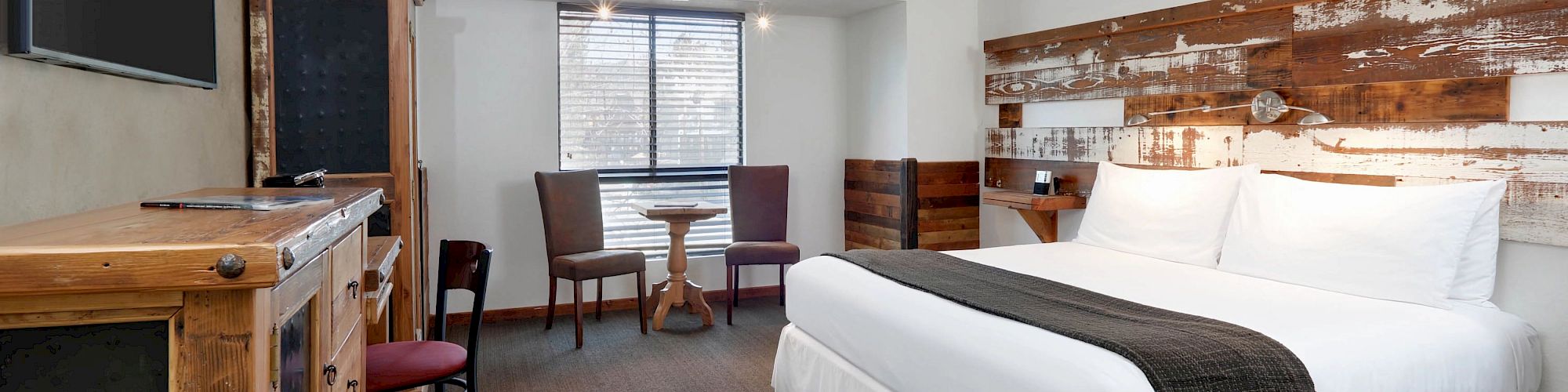 A cozy hotel room with a king-sized bed, rustic wooden furniture, a small round table, two chairs, and a flat-screen TV on the wall.