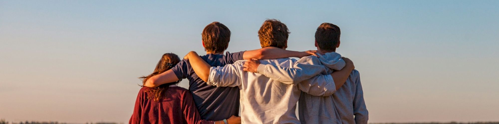 Four people stand in a field with their arms around each other, facing away and looking towards the horizon under a clear sky.