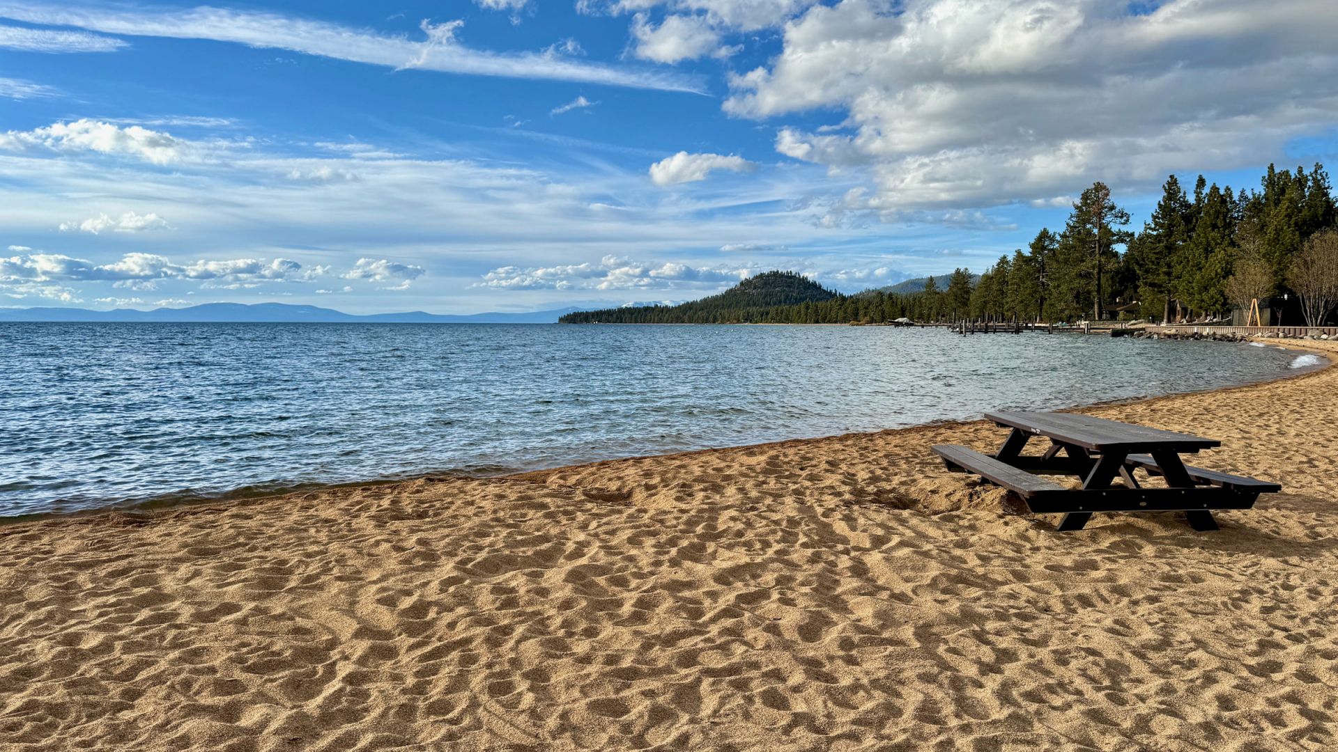 A serene beach scene with golden sand, a lone picnic table, calm blue water, and a partly cloudy sky with distant trees and mountains in the background.