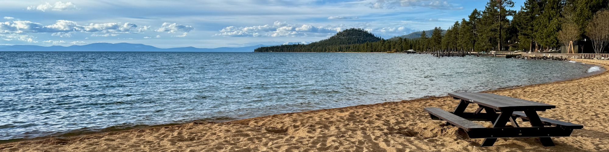 A serene beach scene with golden sand, a lone picnic table, calm blue water, and a partly cloudy sky with distant trees and mountains in the background.