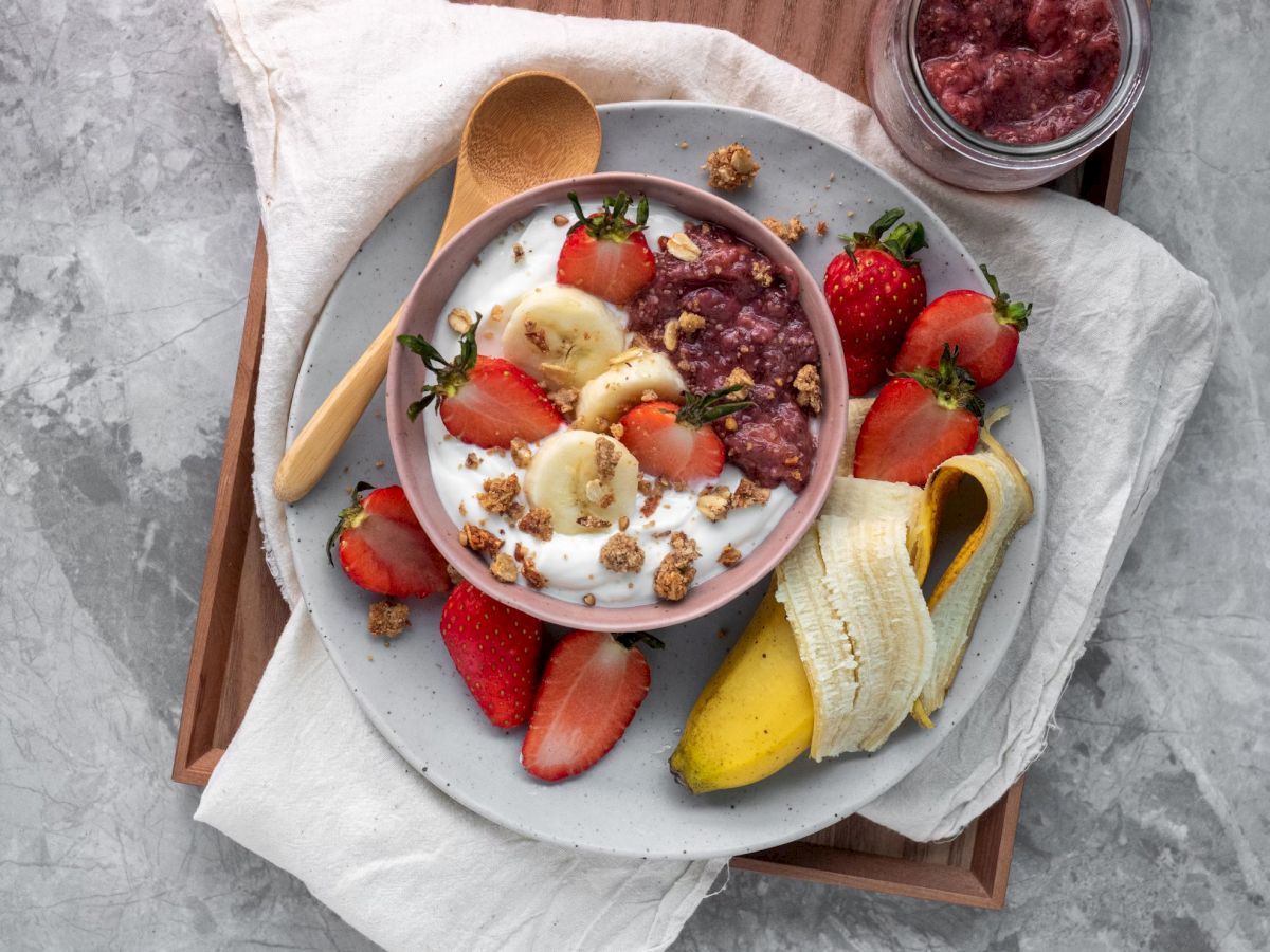 A bowl of yogurt topped with strawberries, bananas, and granola on a tray with more strawberries, a banana, and a jar of spread.