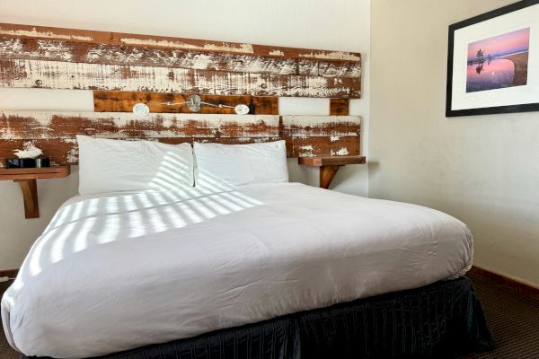 A neatly made, double bed framed by a wooden headboard with two mounted nightstands and a picture on the wall, in a well-lit room.