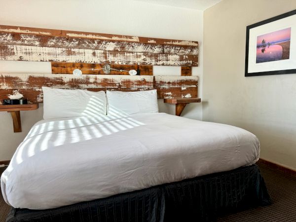 A neatly made, double bed framed by a wooden headboard with two mounted nightstands and a picture on the wall, in a well-lit room.
