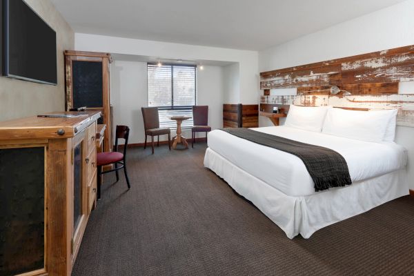 A cozy hotel room with a large bed, rustic wooden furniture, a small table with chairs, and a flat-screen TV on the wall.