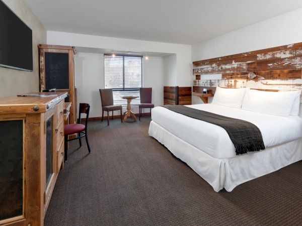 A cozy hotel room with a large bed, rustic wooden furniture, a small table with chairs, and a flat-screen TV on the wall.