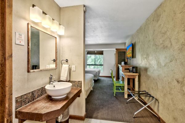 A hotel room includes a sink, mirror, bed, desk, chair, TV, window, and folded luggage rack. The walls are beige, and the carpet is brown.