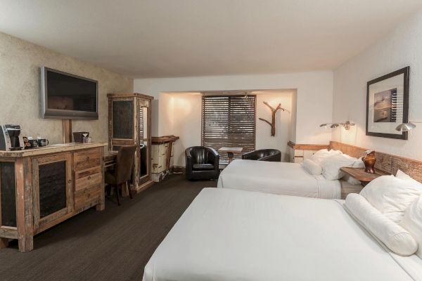 The image shows a cozy hotel room with two queen beds, rustic wooden furniture, a flat-screen TV, and a small seating area near the window.