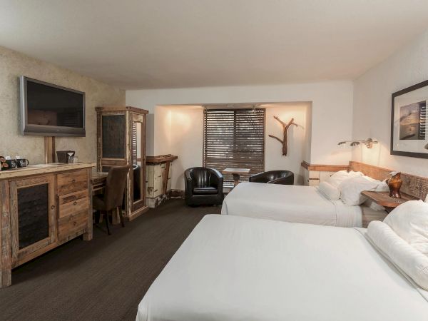The image shows a cozy hotel room with two queen beds, rustic wooden furniture, a flat-screen TV, and a small seating area near the window.