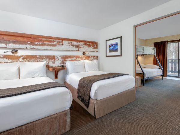 A hotel room featuring two double beds, a bunk bed, neutral decor, wall art, and a balcony with a view of trees outside.