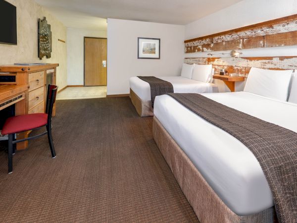 A hotel room with two double beds, a desk with a chair, a flat-screen TV, and wall art décor. The room has a spacious layout and modern furnishings.