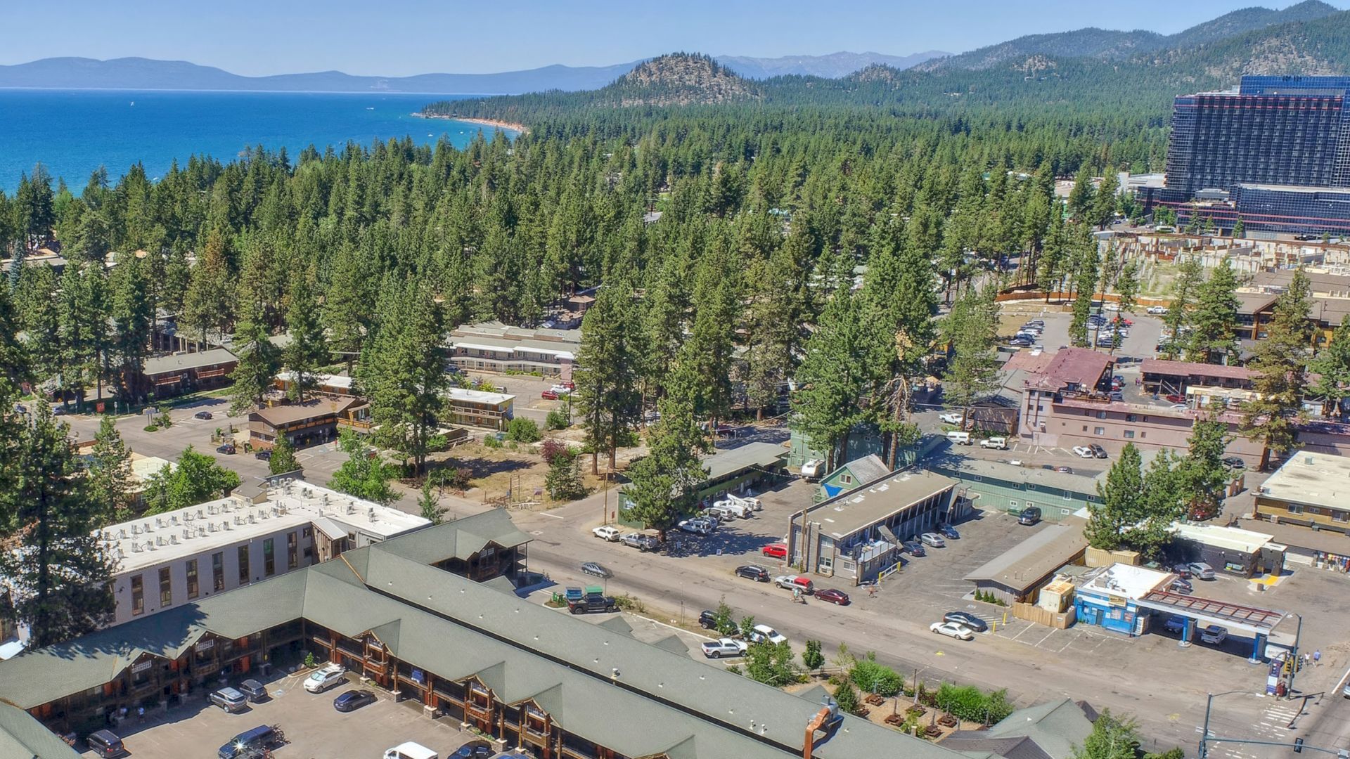 An aerial view of a coastal town with buildings surrounded by trees, adjacent to a large body of water, and mountains in the background.