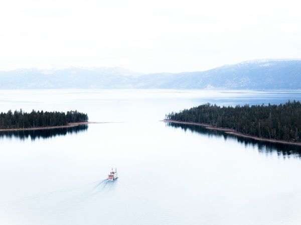 A boat sails between two forested landmasses on a calm lake, surrounded by mountains and an overcast sky in the background.