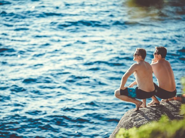Two shirtless individuals are sitting and squatting on a rock by the water, wearing swim shorts and sunglasses, enjoying the peaceful scenery.