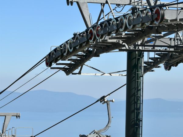 The image shows a gondola lift on a cable, with an ocean or large body of water in the background, and a clear blue sky.