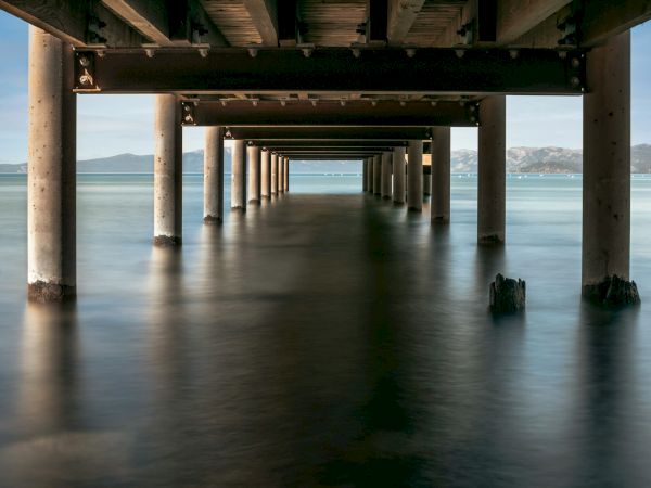 The image shows a view from beneath a wooden pier, with columns extending into calm water, mountains visible in the background.