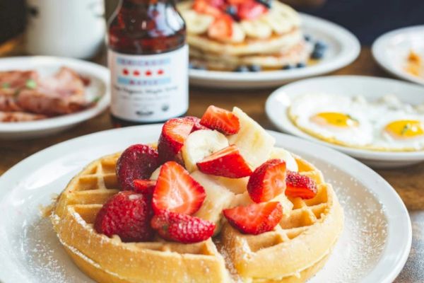 A plate with waffles topped with strawberries and bananas, surrounded by breakfast items like fried eggs, pancakes, and ham.