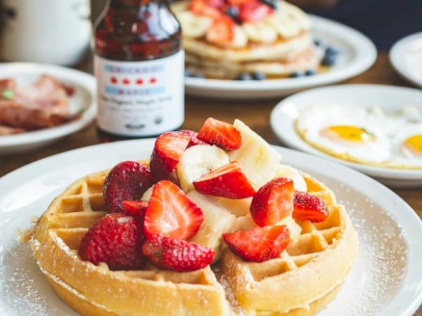 A plate with waffles topped with strawberries and bananas, surrounded by breakfast items like fried eggs, pancakes, and ham.
