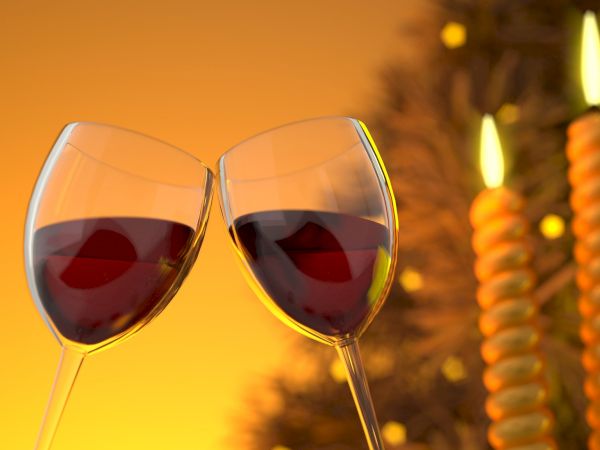 The image shows two wine glasses with red wine clinking together in front of two twisted, lit candles, creating a warm, festive atmosphere.