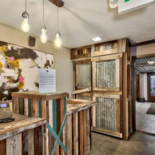 A rustic reception area with a desk, colorful abstract painting, and pendant lights. A hallway with wooden doors leads to another room.