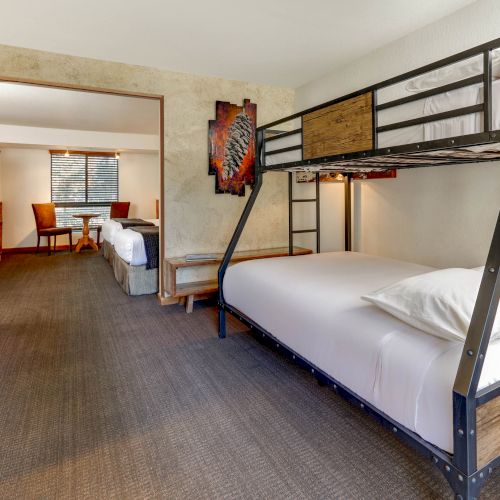 A room with a bunk bed and a single bed, featuring a wooden accent wall, a carpeted floor, and a table with two chairs by the window in the background.