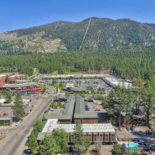 Aerial view of a mountain town with buildings, roads, and parked cars, surrounded by tall trees and a mountainous backdrop ending the sentence.