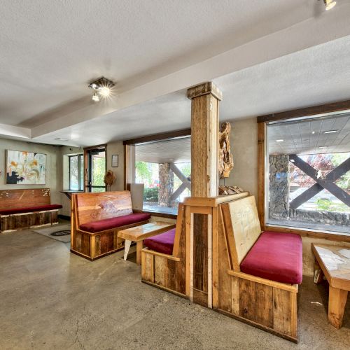 The image shows an indoor waiting area with wooden benches and tables, red cushioned seating, large windows, and art on the walls.