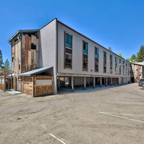 The image shows a multi-story building with a large parking lot in a suburban setting, surrounded by trees and clear skies.