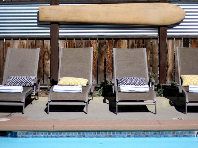 Four chaise lounge chairs sit by a pool with a surfboard above them.
