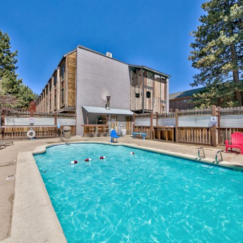 An outdoor swimming pool with lounge chairs, colorful seating, and a building in the background is surrounded by trees and a clear blue sky.