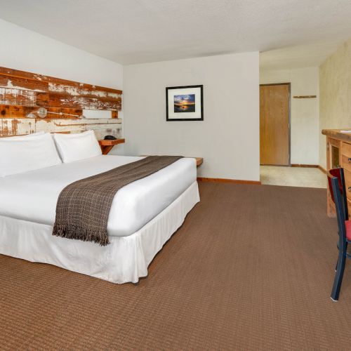 A hotel room with a large bed, a wooden desk, a chair, a wall-mounted TV, and a framed picture on the wall. The decor is rustic.