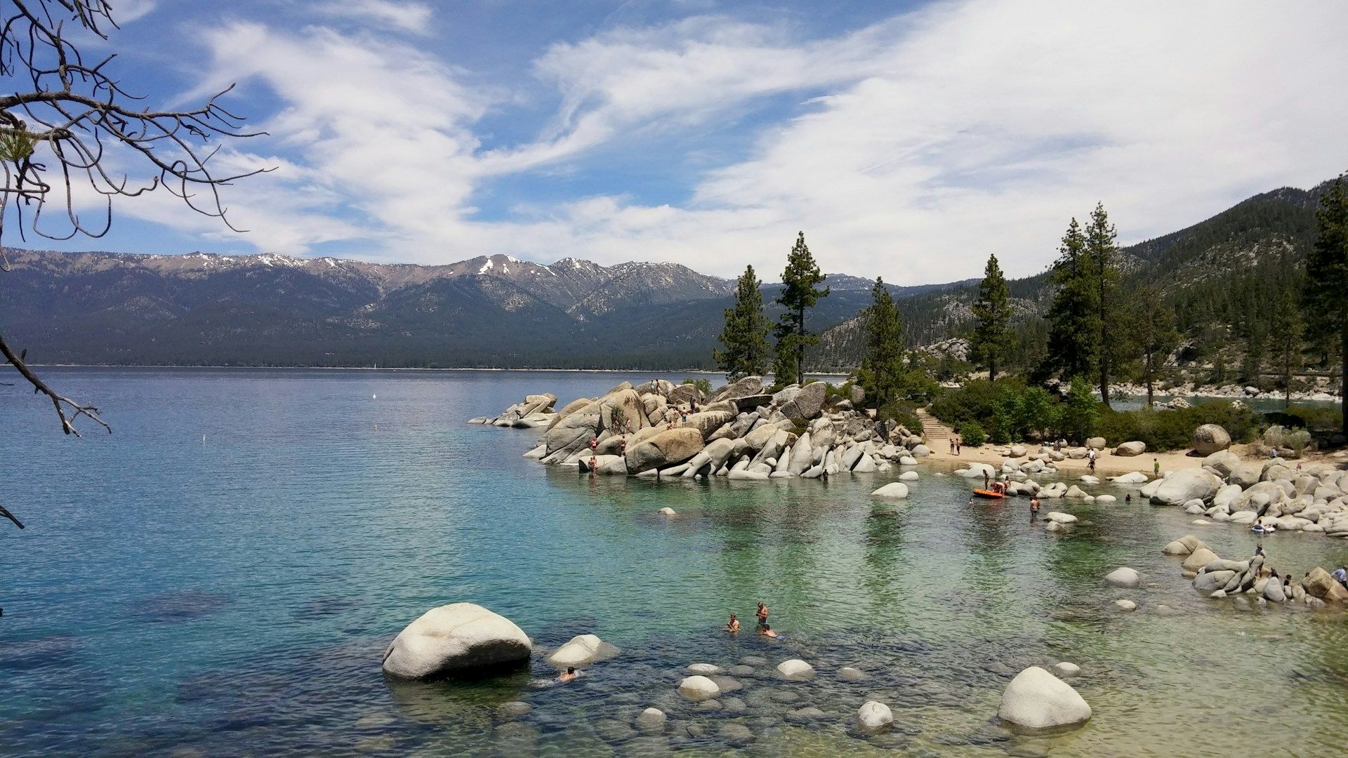 A scenic view of a lake with people swimming and rocks in the foreground, surrounded by trees and mountains in the background.